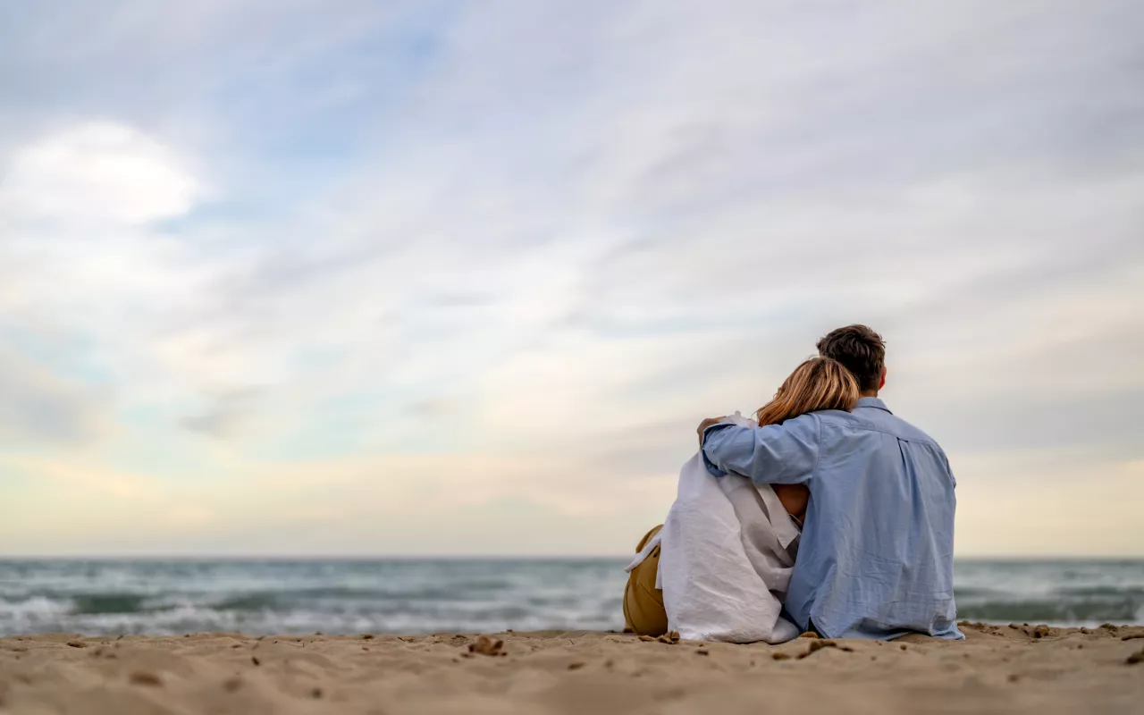 Ein Paar sitzt Arm in Arm am Strand und schaut hinaus auf das Meer. 