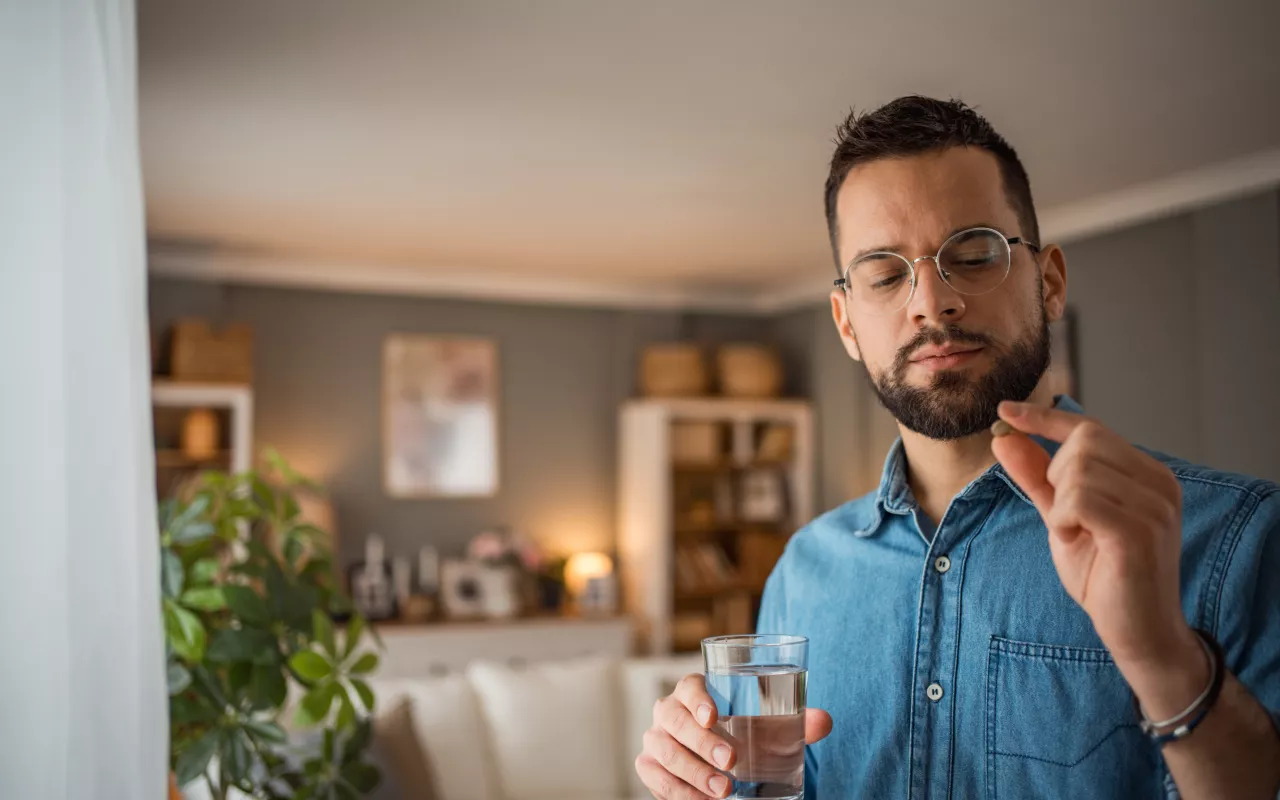 Ein Mann mit braunen Haaren und Bart hält in der einen Hand ein Glas Wasser und in der anderen eine Pille. Diese betrachtet er kritisch.