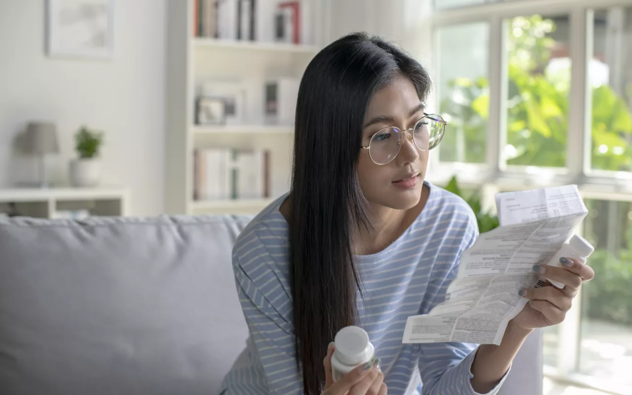 Eine asiatisch gelesene Frau mit Brille sitzt auf einer Couch und hält in der einen Hand eine Medikamentenpackung und in der anderen den Beipackzettel. Sie liest sich den Zettel konzentriert durch.