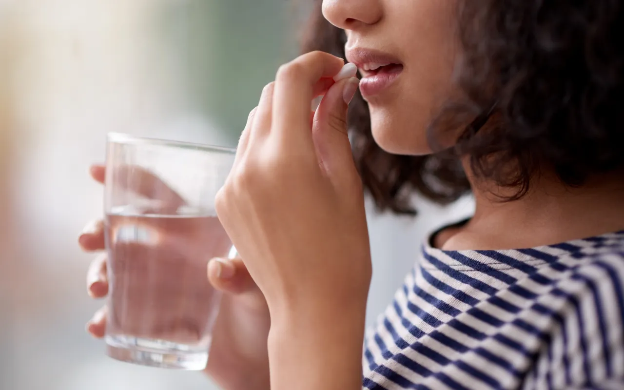 Eine junge Frau mit braunem Haar nimmt eine Tablette ein. Dabei hält sie in der anderen Hand ein Glas Wasser.
