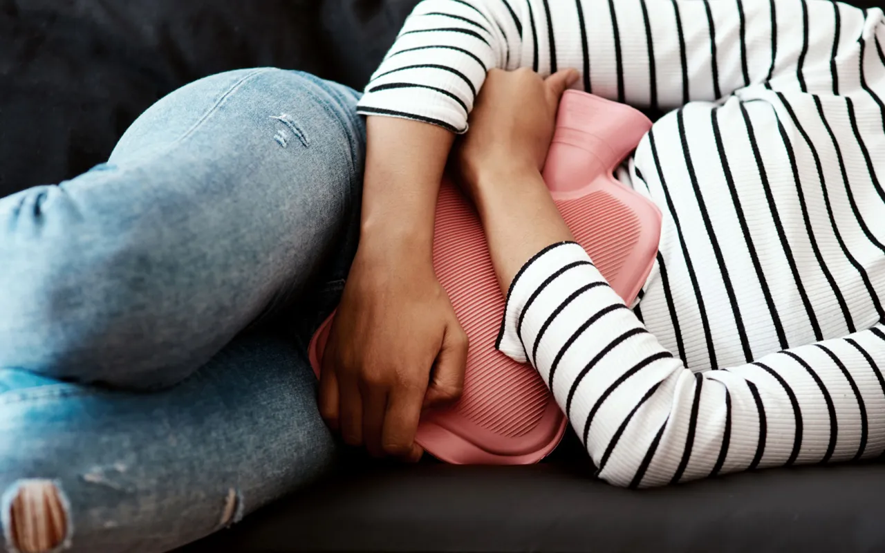 Beschnittene Aufnahme einer Frau, die zu Hause auf dem Sofa eine Wärmflasche gegen den Bauch hält