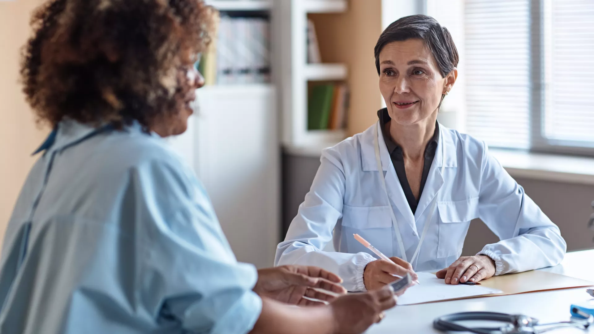 Es sind zwei Frauen zu sehen, die an einem Schreibtischsitzen. Die eine Frau ist eine Ärztin und erklärt der vemeintlichen Patientin etwas. Beide wirken entspannt und interessiert. 