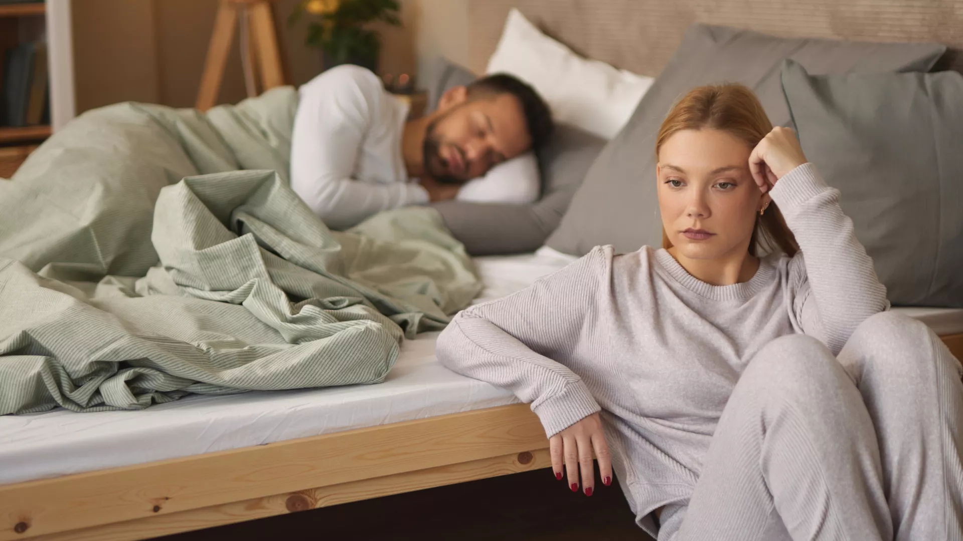 Eine blonde Frau sitzt neben einem Bett und lehnt sich an die Seite davon. Ein Mann schläft im Hintergrund in diesem Bett. Ihr Blick ist nachdenklich und pessimistisch.