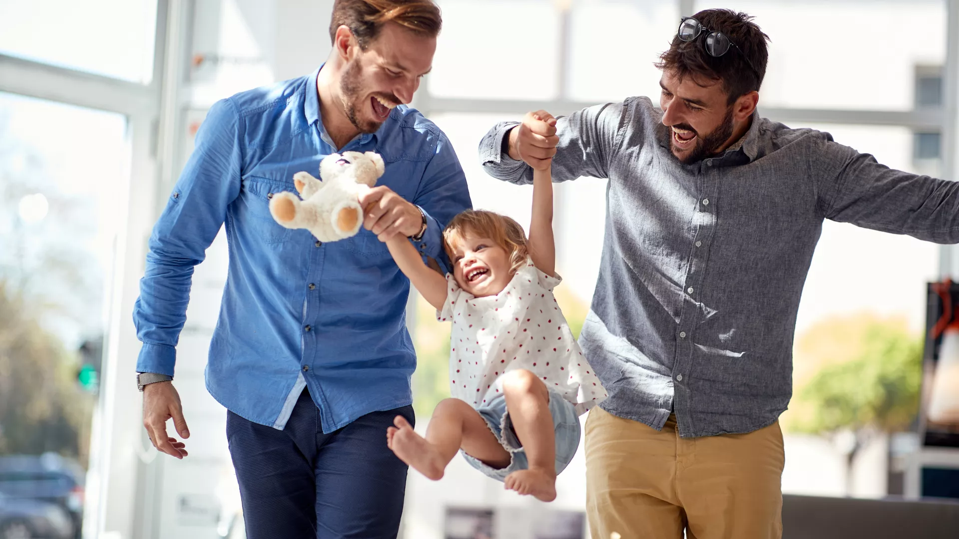 Zwei Männer lächeln und halten an ihren Händen ein kleines Mädchen(ihre gemeinsame Tochter) und heben sie gemeinsam in die Luft. Alle lachen dabei beherzt und sehen glücklich aus.