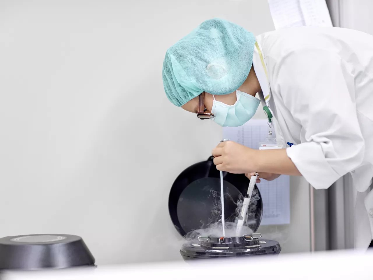 Eine Person mit Mundschutz, Haube und Weißem Kittel ist über einen Atickstoff-Kühlbehälter gebeugt und entnimmt dort eingefrorene Proben. Dabei hält sich zwei Hilfmittel in der Hand, um den kalten Behälter nicht berühren zu müssen. Kalter Dampf steigt aus dem Behälter empor.