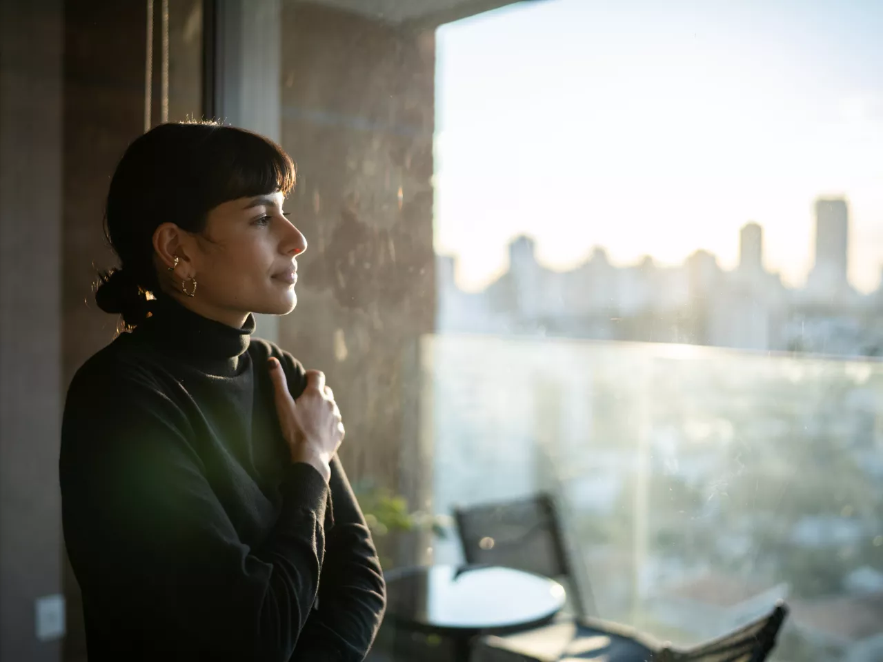 Eine Frau mit braunem Haar und Pony schaut gedankenverloren aus dem Fenster. Das Licht scheint sanft herein und die Skyline einer Stadt ist verschwommen zu erkennen.