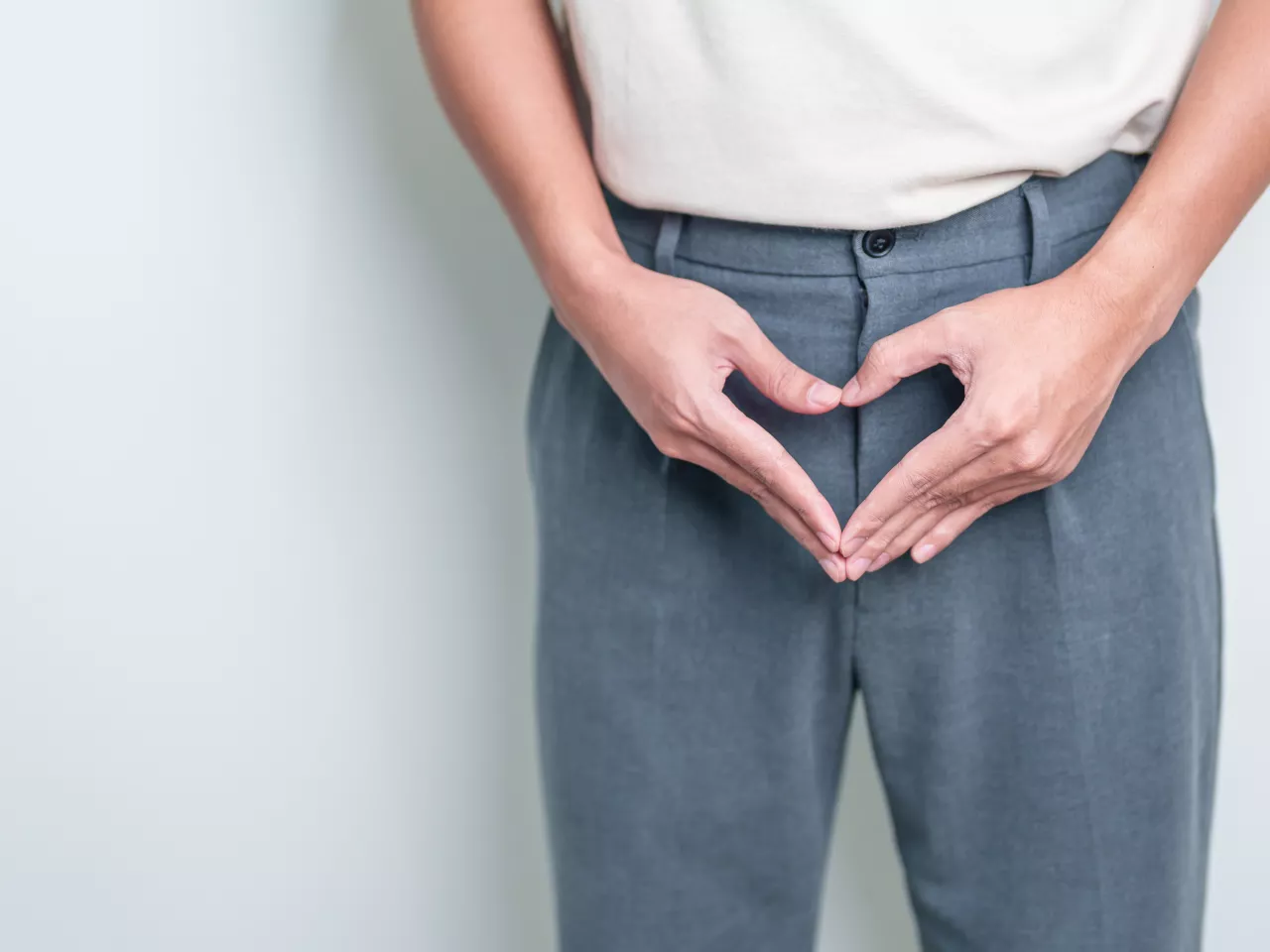 Die Körpermitte eines Mannes in grauer Hose und weißem Shirt wird gezeigt. Der Rest des Körpers ist nicht zu sehen. Der Mann hält seine Hände vor seinem Schritt.