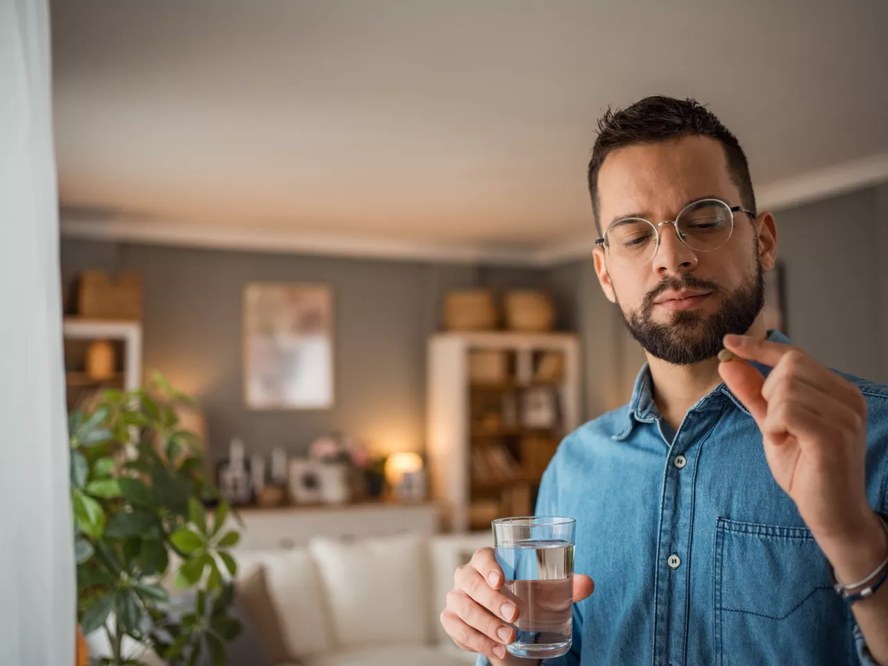 Ein Mann mit braunen Haaren und Bart hält in der einen Hand ein Glas Wasser und in der anderen eine Pille. Diese betrachtet er kritisch.