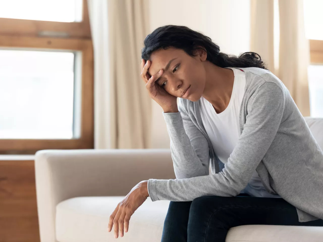 Eine schwarze Frau sitzt auf einem Sofa und hat ihren Kopf auf ihrer Hand abgestützt. Sie schaut bedrückt und nachdenklich auf den Boden.