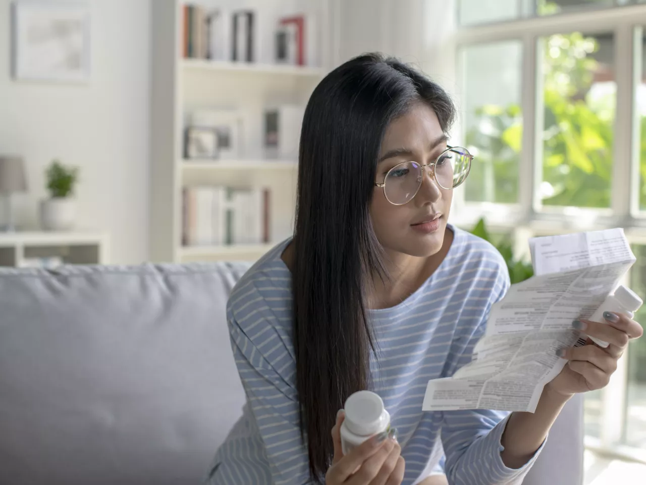 Eine asiatisch gelesene Frau mit Brille sitzt auf einer Couch und hält in der einen Hand eine Medikamentenpackung und in der anderen den Beipackzettel. Sie liest sich den Zettel konzentriert durch.