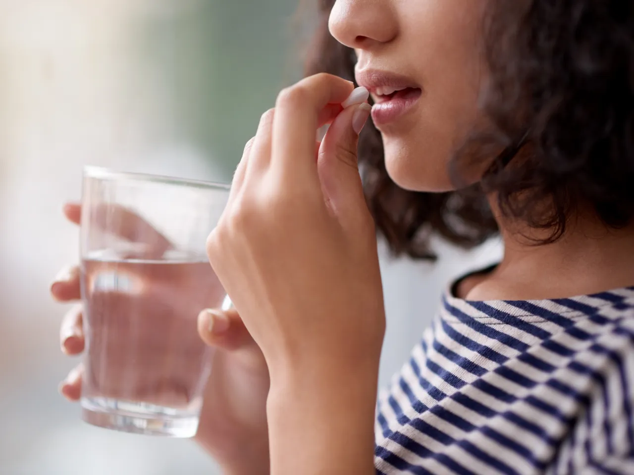 Eine junge Frau mit braunem Haar nimmt eine Tablette ein. Dabei hält sie in der anderen Hand ein Glas Wasser.