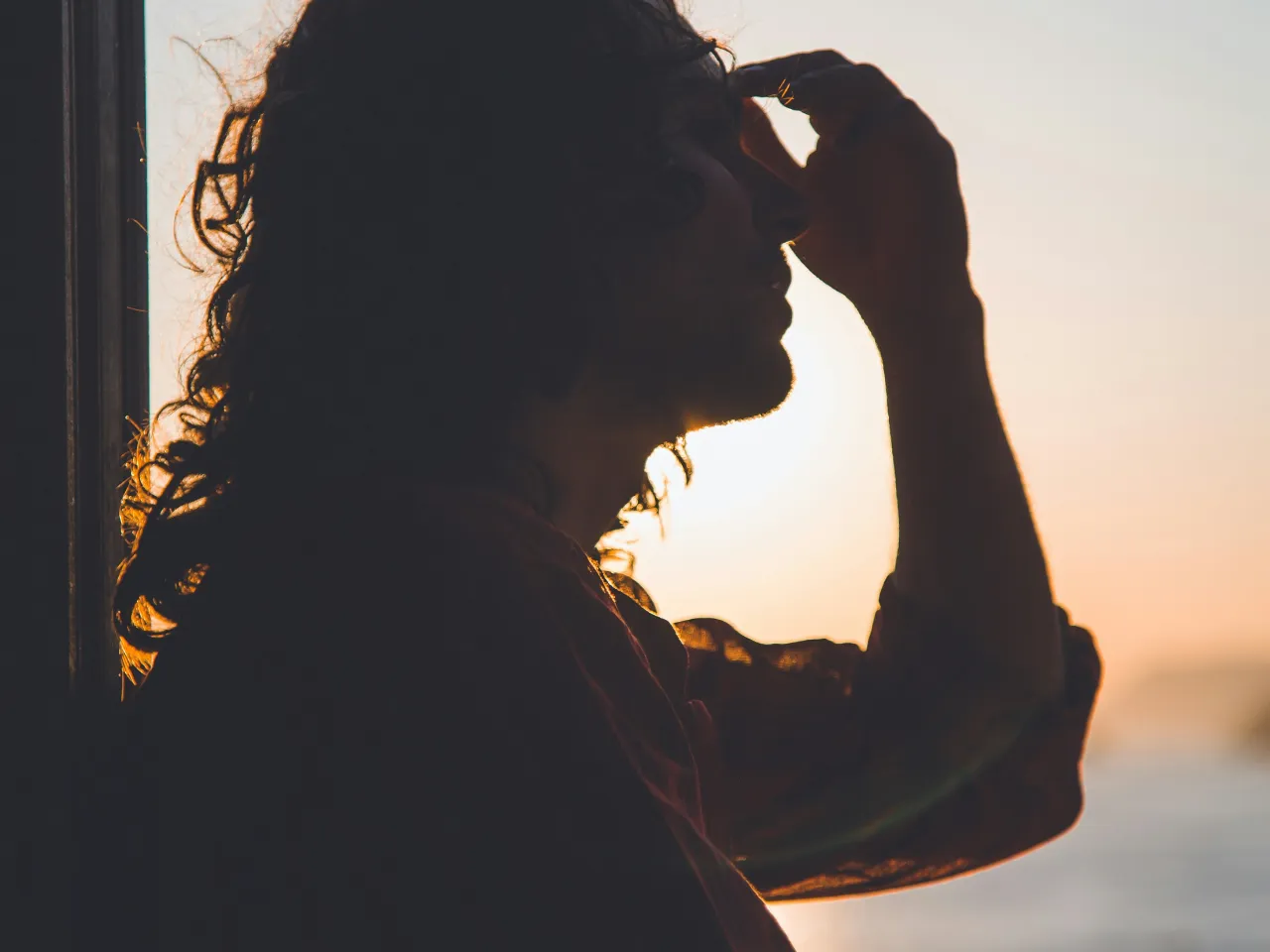 Frau sitzt vor einem Fenster, die Sonne scheint hinen. Sie hält sich die Stirn, wegen Kopfschmerzen.